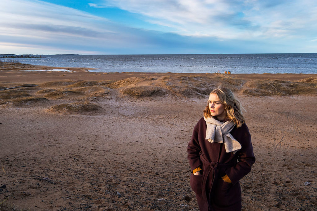 Hanna Parhaniemi seisoo hiekkarannalla, takki päällä, hieman sivusta kuvattuna.